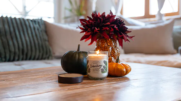 Brennende Zirbenkerze auf einem Holztisch, umgeben von Herbstdekoration wie K&uuml;rbissen und einer roten Blume.