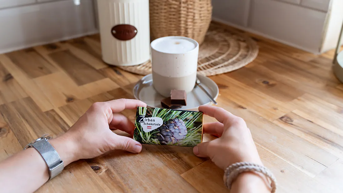 Person h&auml;lt eine Packung Zirbenschokolade &uuml;ber einem Holztisch, neben einer Tasse auf einem Untersetzer.